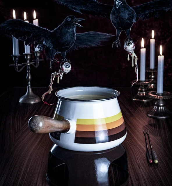 Still life with a pot, raven figurines holding eyeballs, candles in brass candlesticks on a wooden table against a dark backdrop.