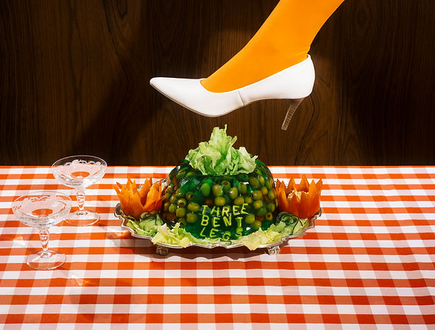 Woman's foot in a white high heel steps on a green gelatin dessert on a checkered tablecloth with wooden background.