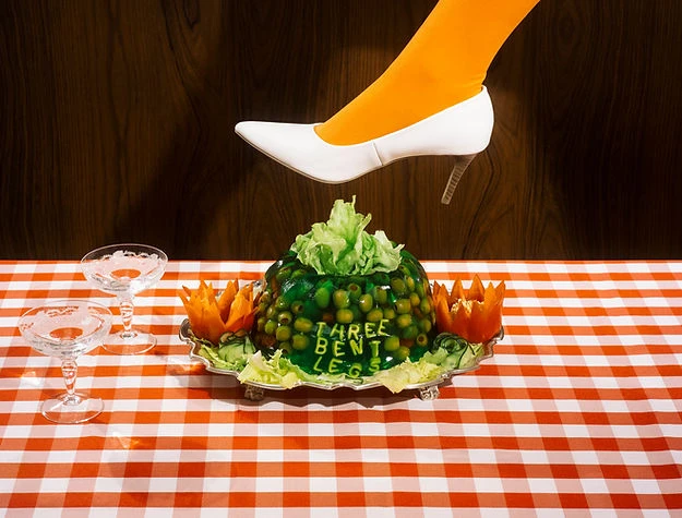 Woman's foot in a white high heel steps on a green gelatin dessert on a checkered tablecloth with wooden background.