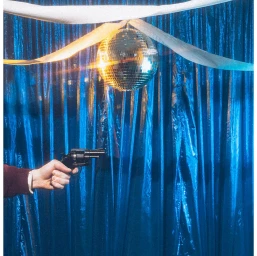 Hand holding a revolver against a blue tinsel curtain beneath a mirrored disco ball with white ribbons