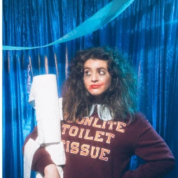A woman holding a roll of toilet paper in front of a blue tinsel curtain, photographed by Three Bent Legs.