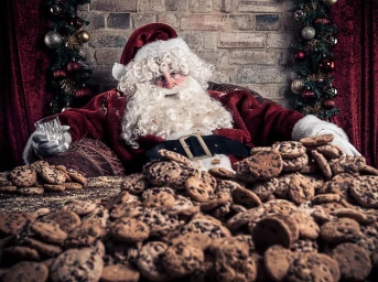 Santa Claus wearing red coat and white beard sitting behind a massive pile of chocolate chip cookies holding a glass