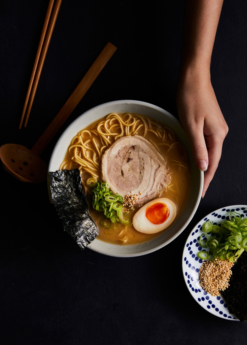 Overhead shot of a bowl of ramen with noodles, pork, egg, and seaweed, held by a hand, with another plate of ingredients nearby.