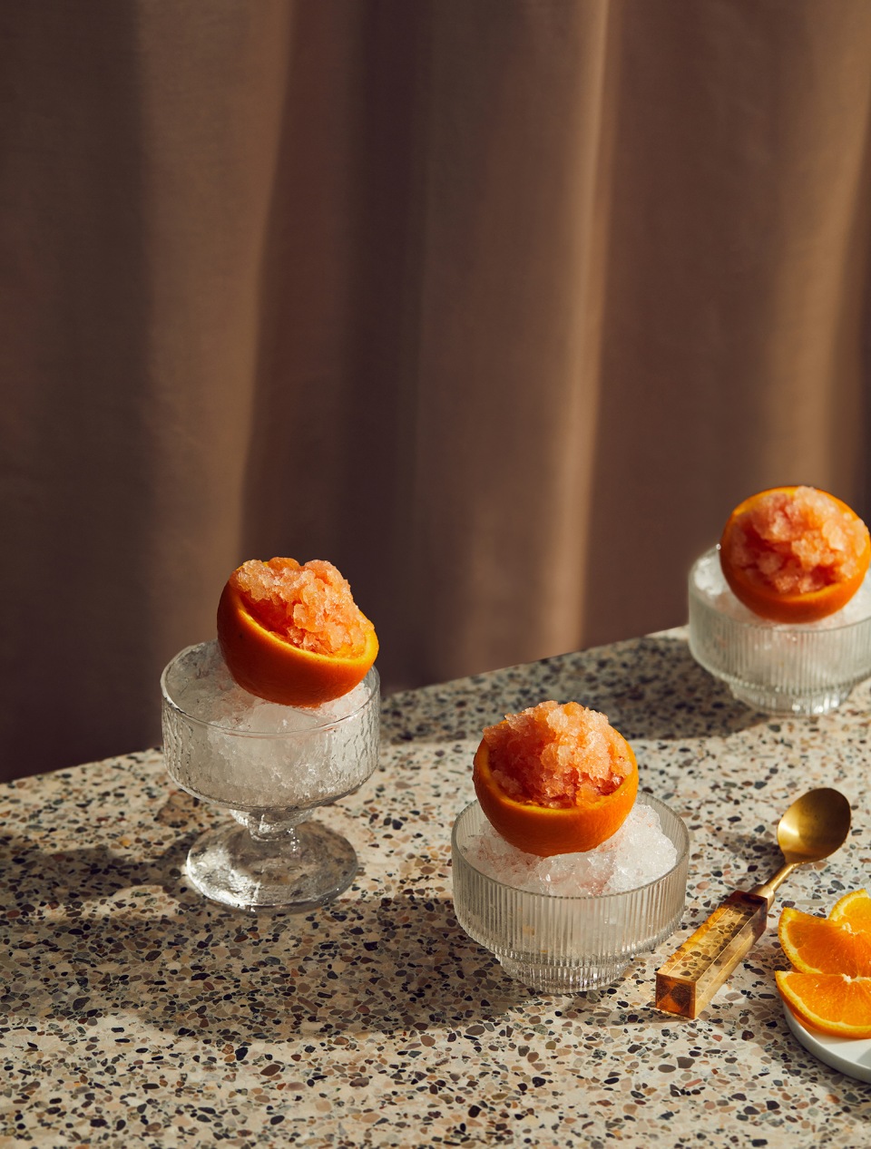 Three grapefruit granitas served in halved grapefruits on crushed ice in glass bowls, displayed on a speckled tabletop.