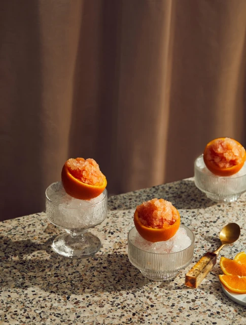 Three grapefruit granitas served in halved grapefruits on crushed ice in glass bowls, displayed on a speckled tabletop.