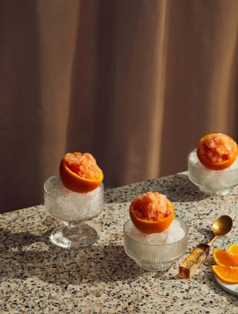 Three grapefruit granitas served in halved grapefruits on crushed ice in glass bowls, displayed on a speckled tabletop.