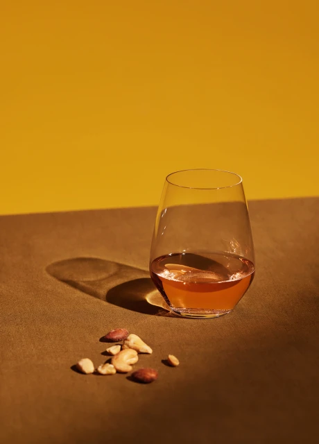 Still life shot of a glass of amber liquid and a small pile of mixed nuts on a brown surface against a yellow background. 