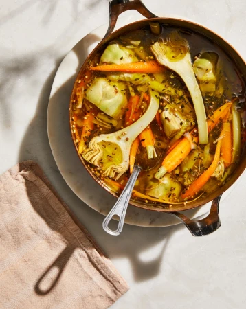 Overhead view of a copper pot filled with artichoke hearts and carrot broth soup, garnished with herbs and served on a marble surface.