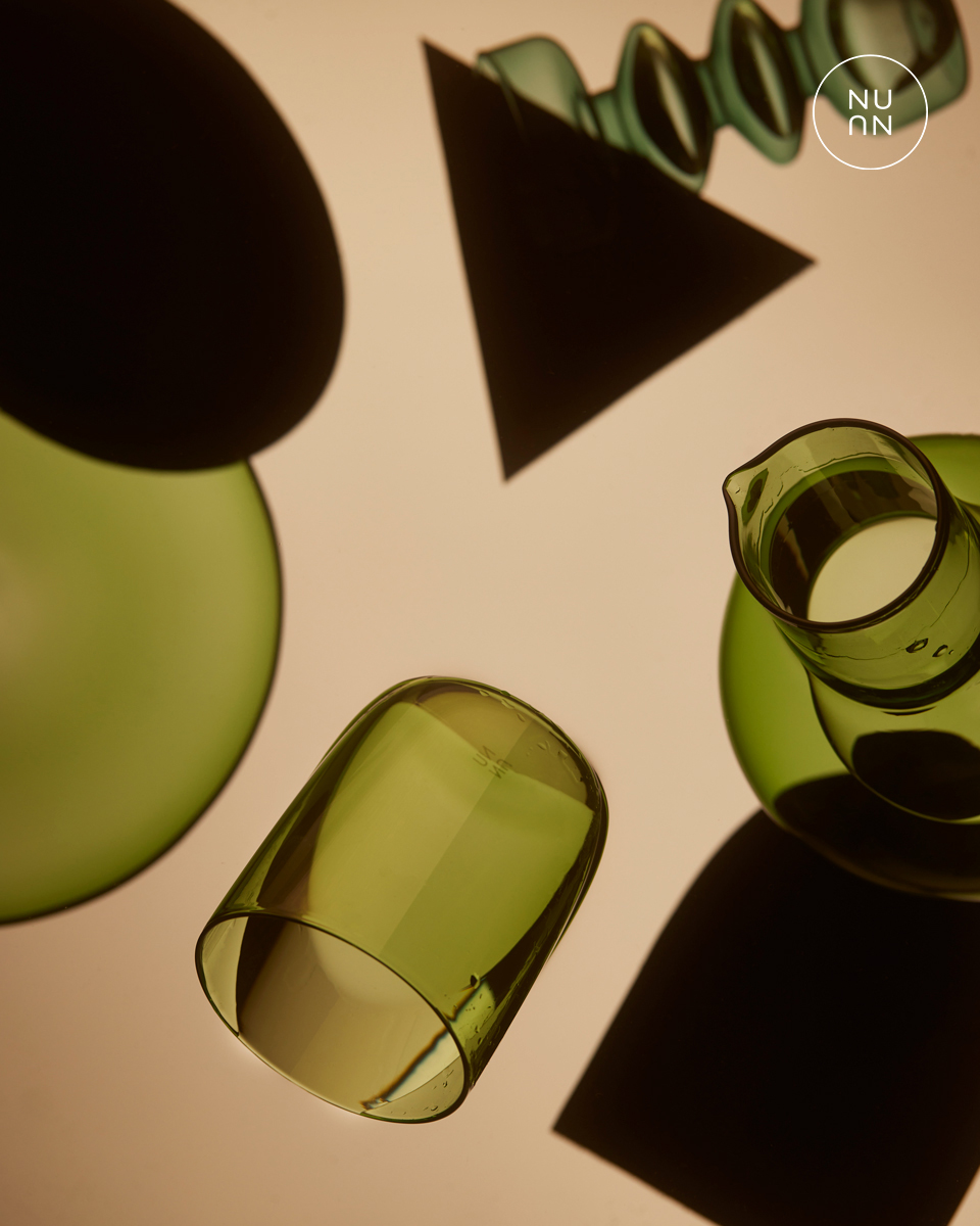 Overhead studio shot of green-tinted glass drinkware including cups and a pitcher, with geometric shapes casting shadows on a beige surface.