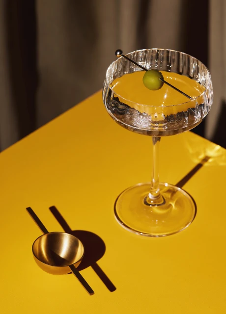 Elegant cocktail in crystal glass on yellow table with olive garnish, highlighting a sophisticated and stylish drink presentation.