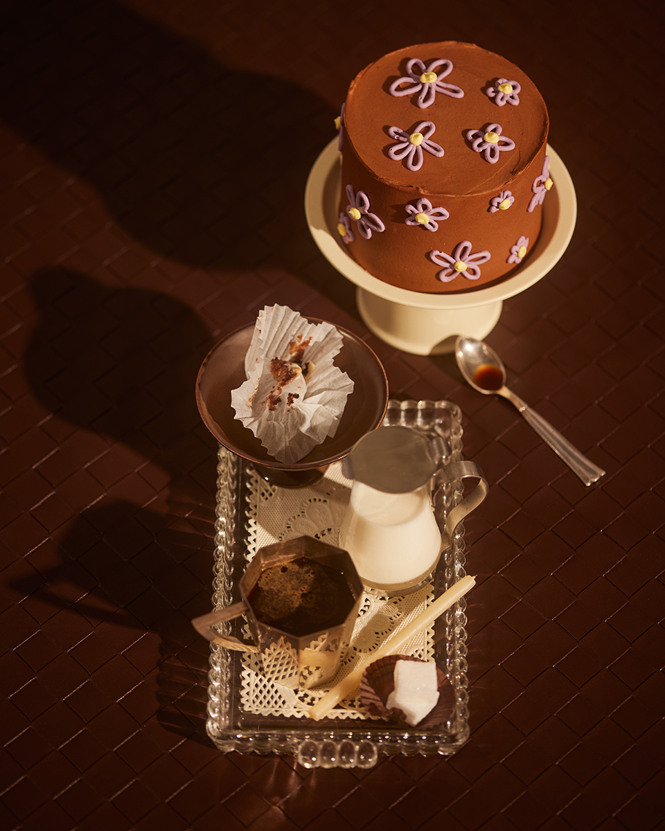 Chocolate cake with flower decorations on a cake stand, alongside a coffee, milk, and chocolates arranged on a decorative glass tray.
