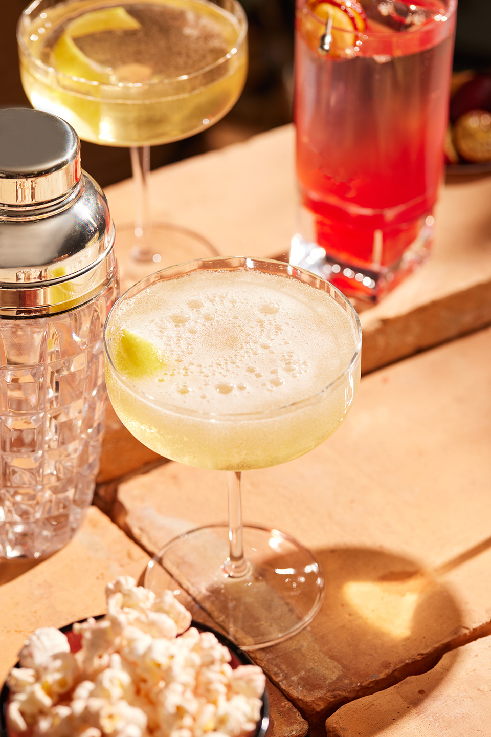 A variety of cocktails in stemmed glasses and a shaker, next to a bowl of popcorn, sit on a textured surface with bright, directional lighting.
