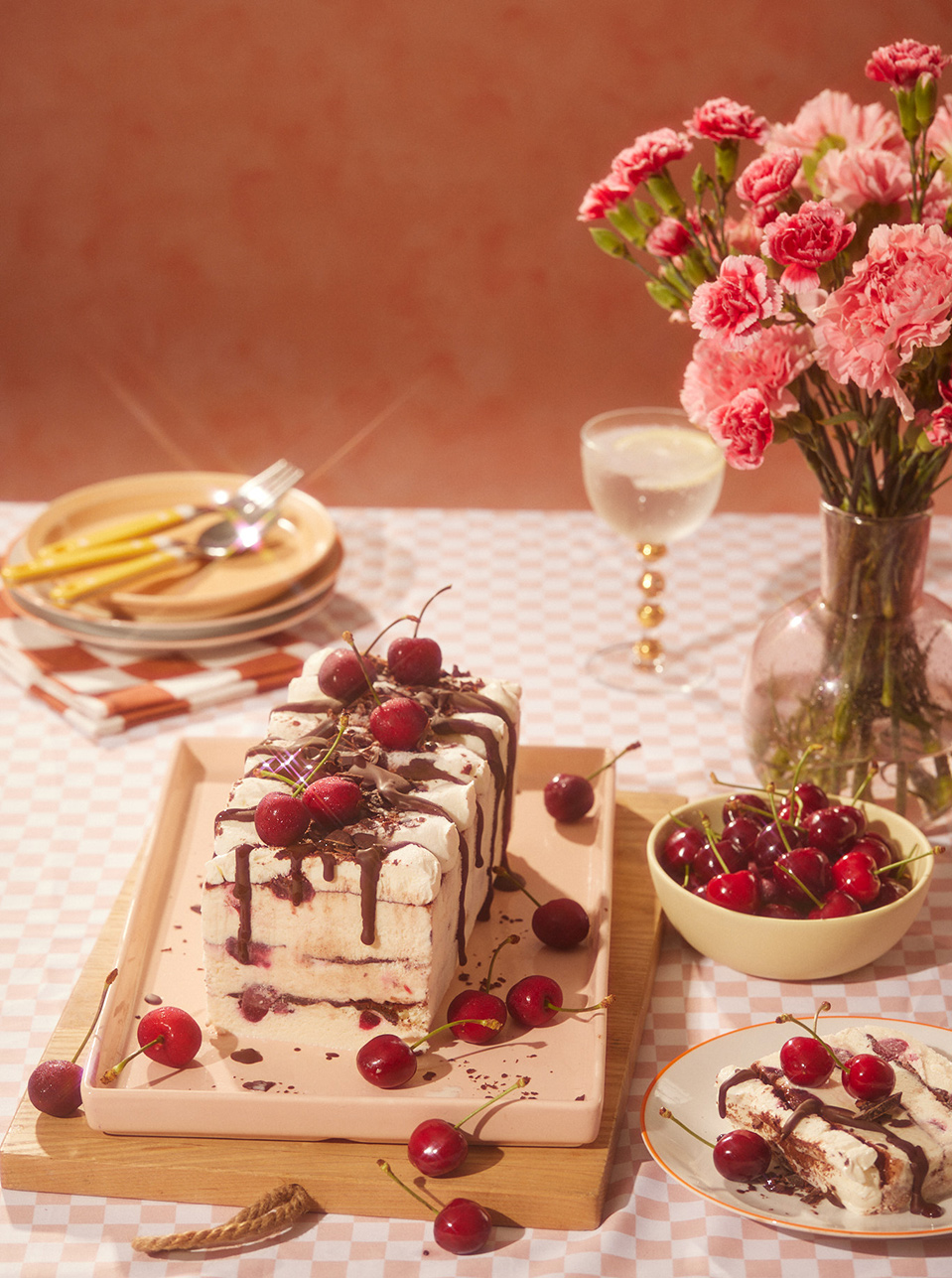 Still life photo of cherry dessert loaf topped with chocolate, alongside fresh cherries, plates, and a glass on a checkered tablecloth.