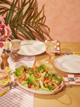 Overhead shot of shrimp salad on an oval platter, pink cocktails, white plates, and pink flowers on a table with a textured pink wall.