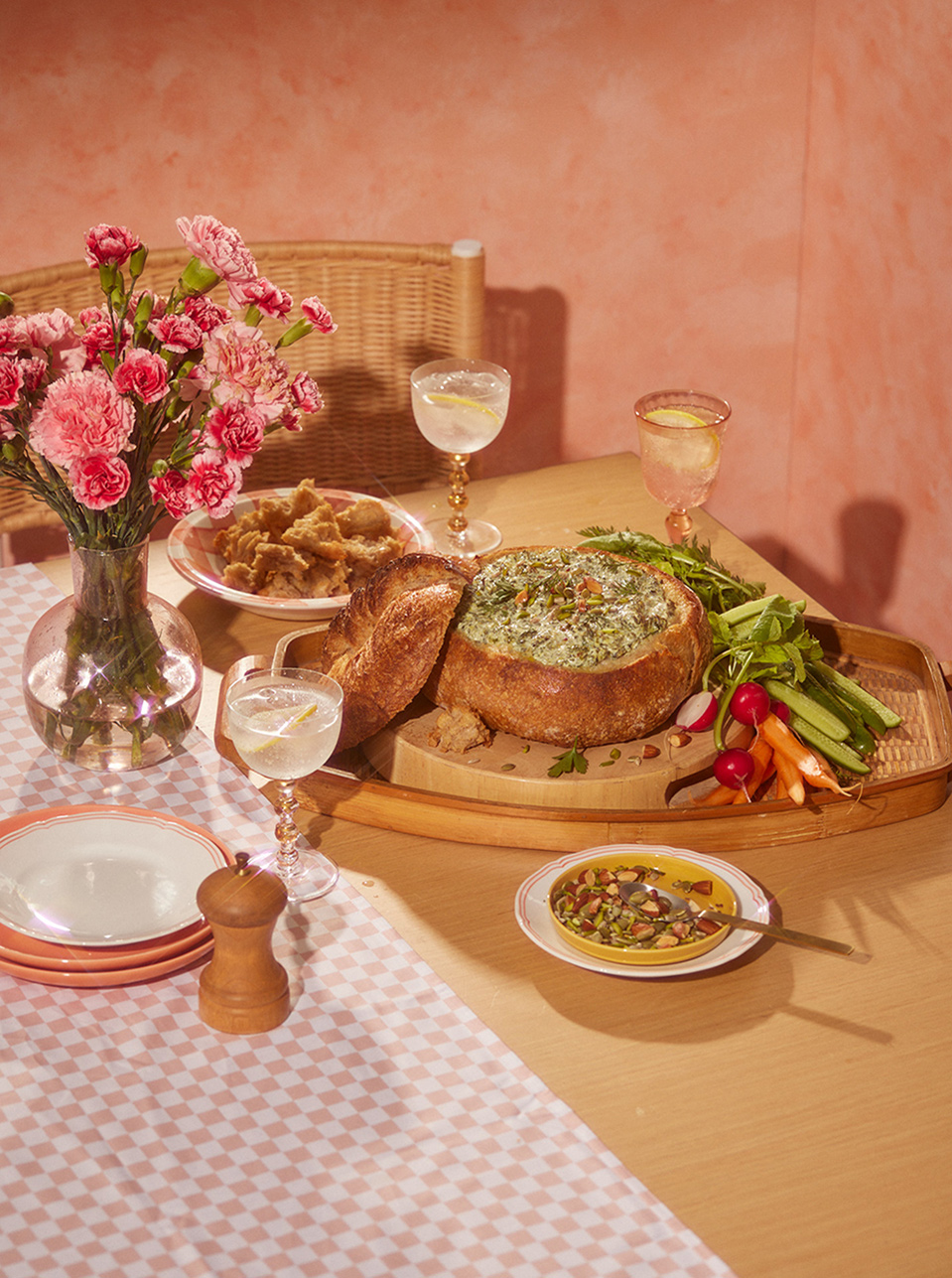 Overhead shot of a table laden with food and drinks, featuring a bread bowl dip, appetizer, flowers, and beverages in a cozy indoor setting.