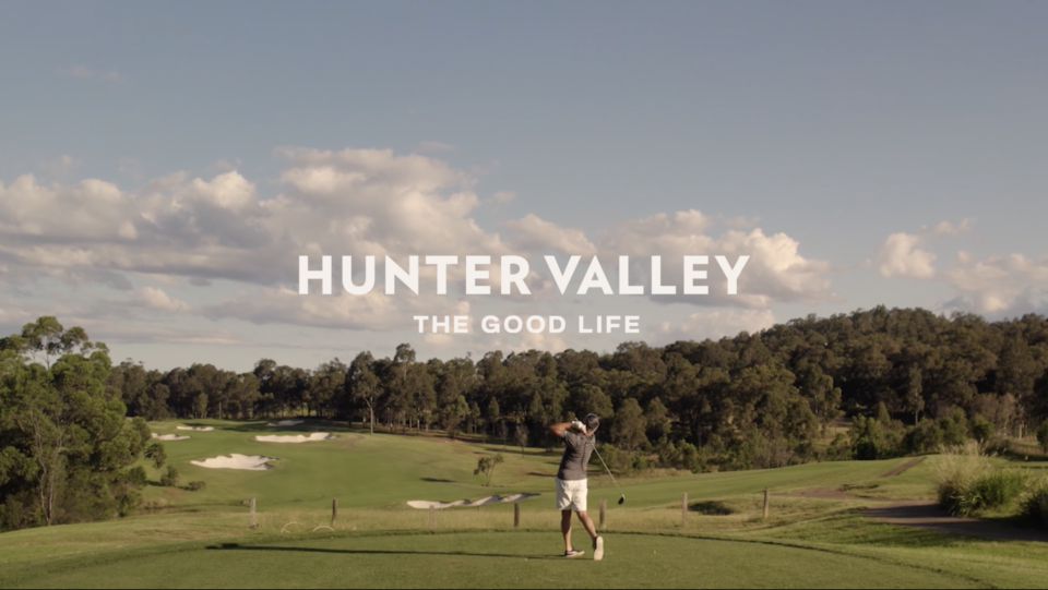 Golfer teeing off on a green golf course in Hunter Valley under a partly cloudy sky, trees in the background.