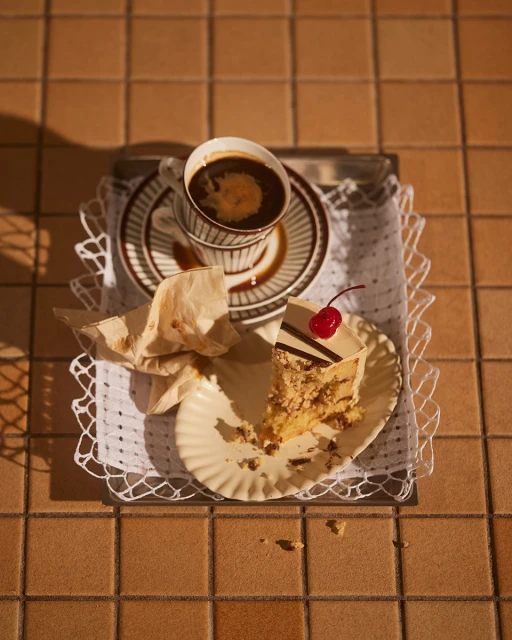Slice of cake with a cherry on a plate and cup of coffee on a silver tray, brown and white color scheme, lit by sunlight.