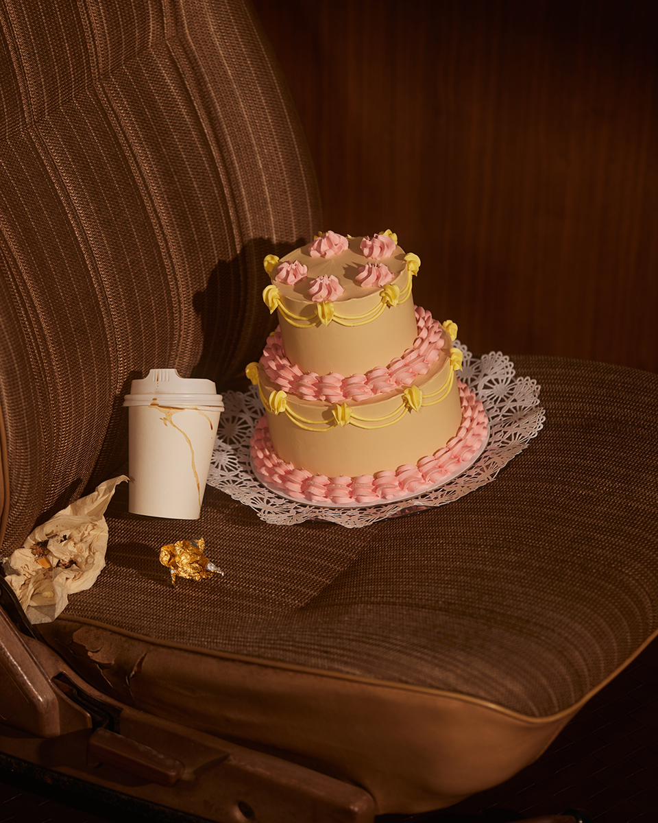 Two-tier cake with pink frosting sits on a lace doily alongside a coffee cup and candy wrapper inside of a vehicle.