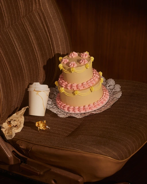 Two-tier cake with pink frosting sits on a lace doily alongside a coffee cup and candy wrapper inside of a vehicle.