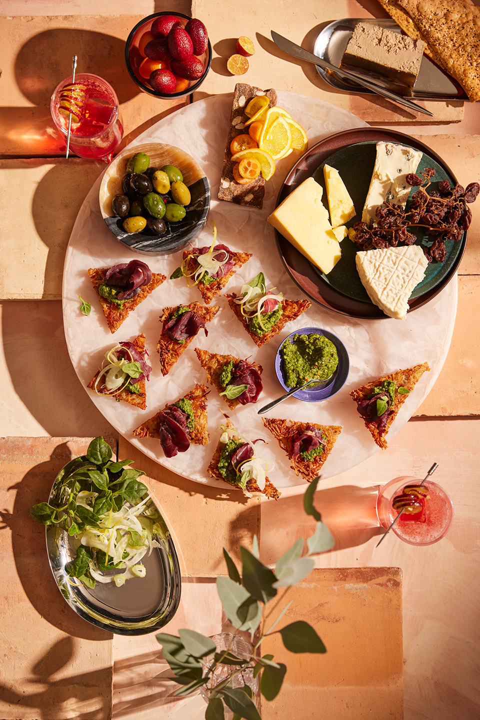 Overhead shot of an assortment of gourmet appetizers including toasts, cheeses, olives, fruit, and cocktails on a textured surface.