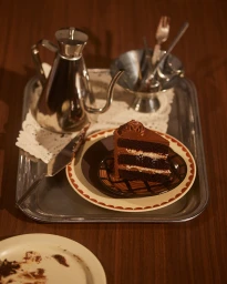 A slice of layered chocolate cake on patterned plates resting on a large silver tray with a metal kettle and silverware.