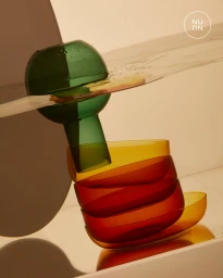 Stacked coloured glassware including green glass bottle and amber and red bowls on reflective surface with water splashes.