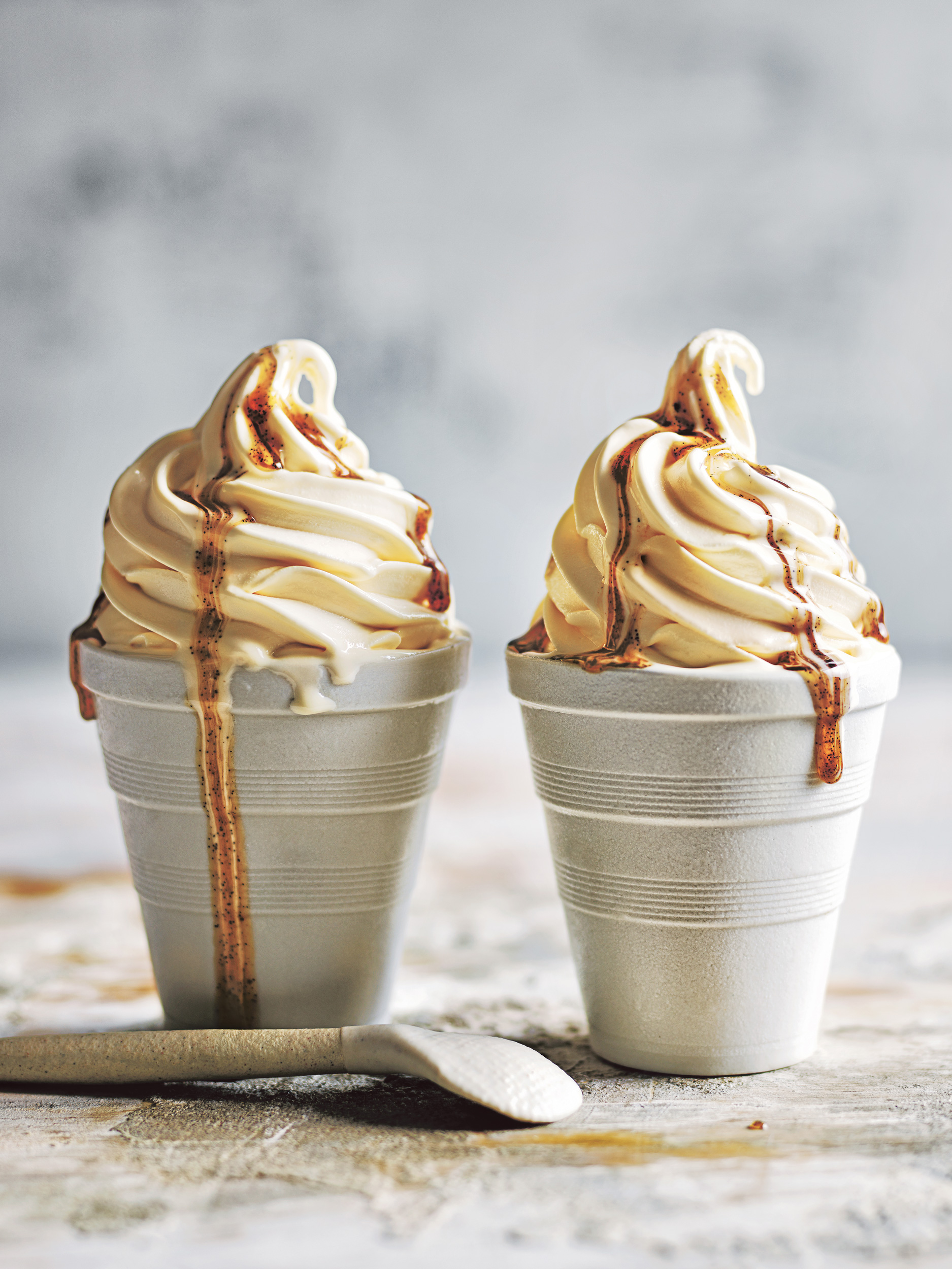 Two servings of soft serve ice cream in paper cups with caramel drizzle sit on a textured surface, illuminated by bright lighting.