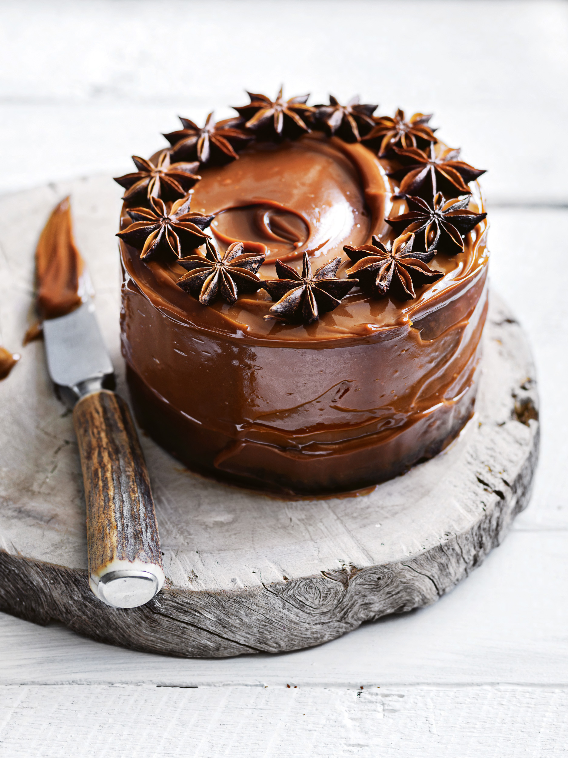 Overhead close-up of a caramel cake topped with star anise, on a wood slice, with a knife, sitting on a white wood surface.