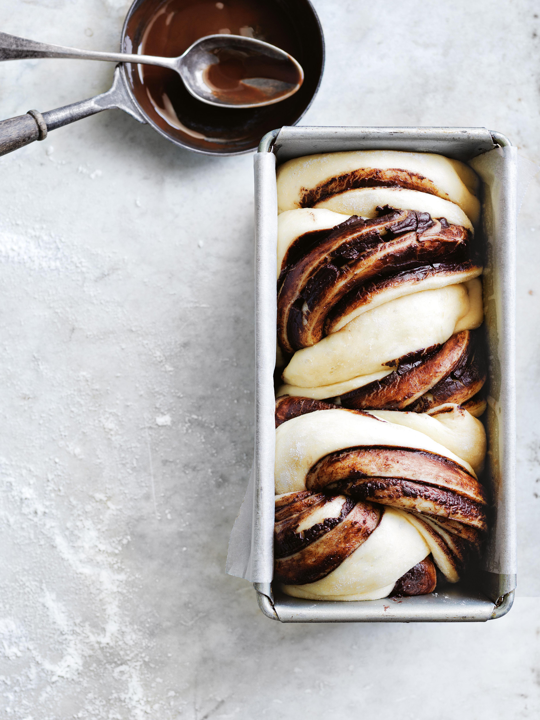 Close-up of a twisted chocolate babka in a metal loaf pan, next to a bowl of melted chocolate and a spoon, on a light background.