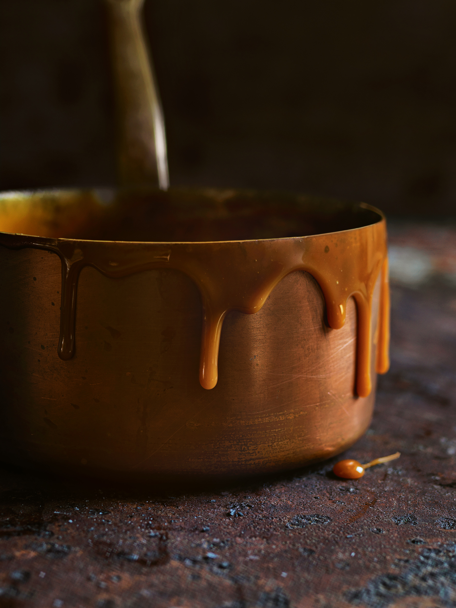 Copper pot with dripping caramel sauce on a dark, textured surface. Culinary still life showcases warm tones and rich texture.