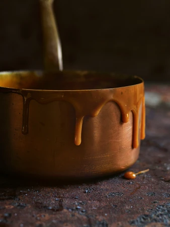 Copper pot with dripping caramel sauce on a dark, textured surface. Culinary still life showcases warm tones and rich texture.