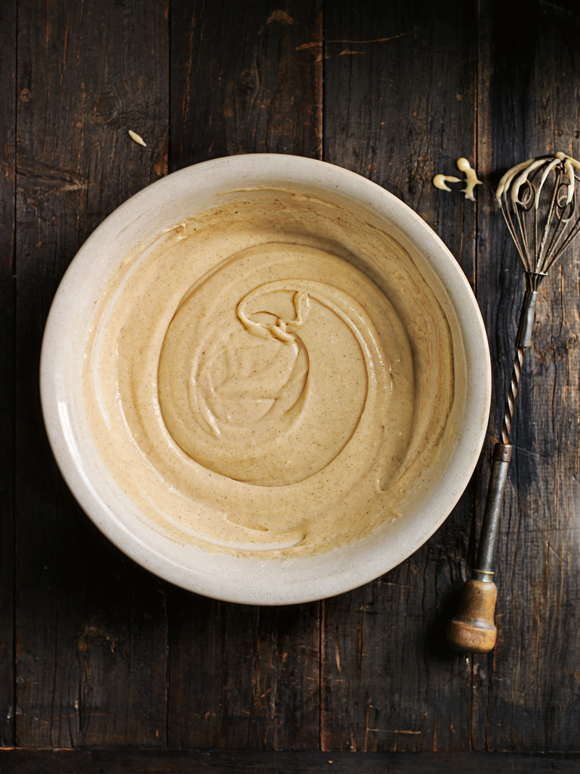 Bowl of batter on a dark wood surface, next to a metal whisk. Food photography showcasing ingredients and cooking preparation.