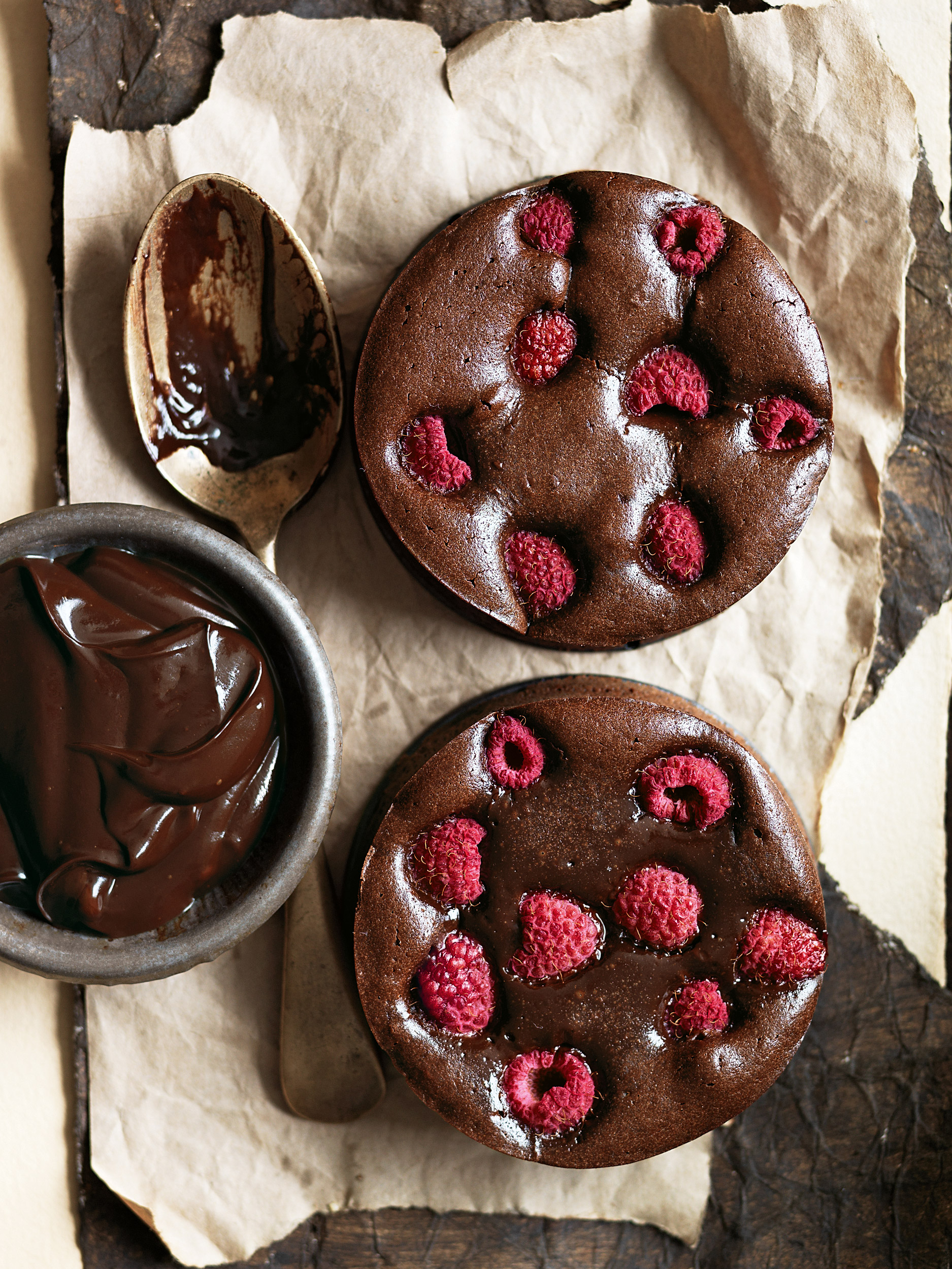 Two chocolate raspberry cakes on parchment paper next to a bowl of chocolate sauce and a spoon with chocolate.