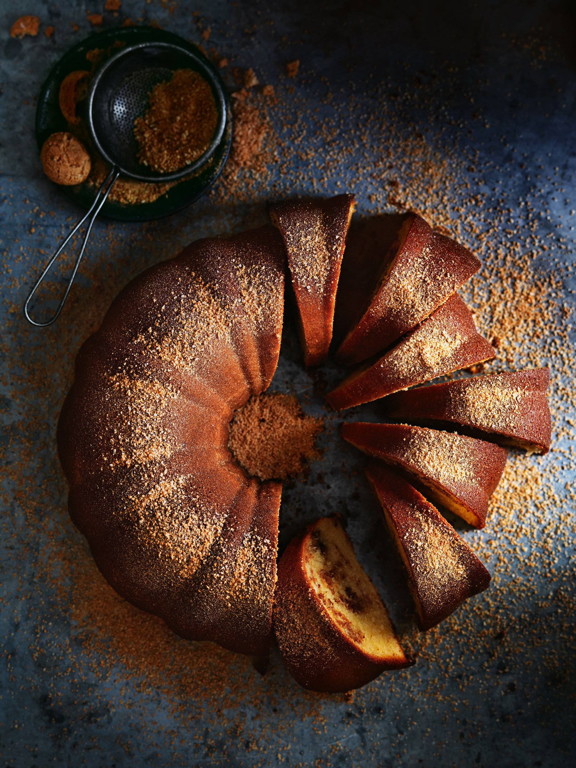 Overhead shot of sliced Bundt cake with dusting powder, placed on a textured blue surface with a sieve and decorative plate.