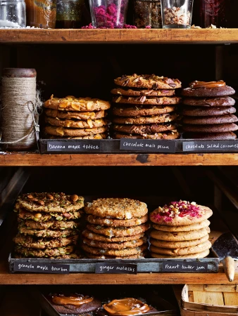 Stacked cookies with different toppings on rustic wooden shelves. Variety of flavors like maple toffee, chocolate pecan, and peanut butter.