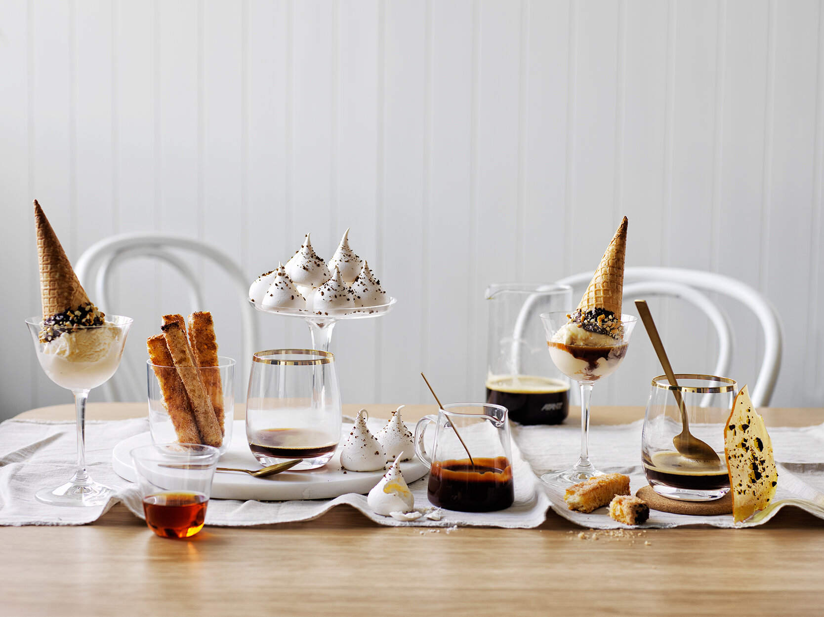 Affogato desserts with ice cream in cones and small meringue desserts are arranged on a wooden table with white chairs behind.