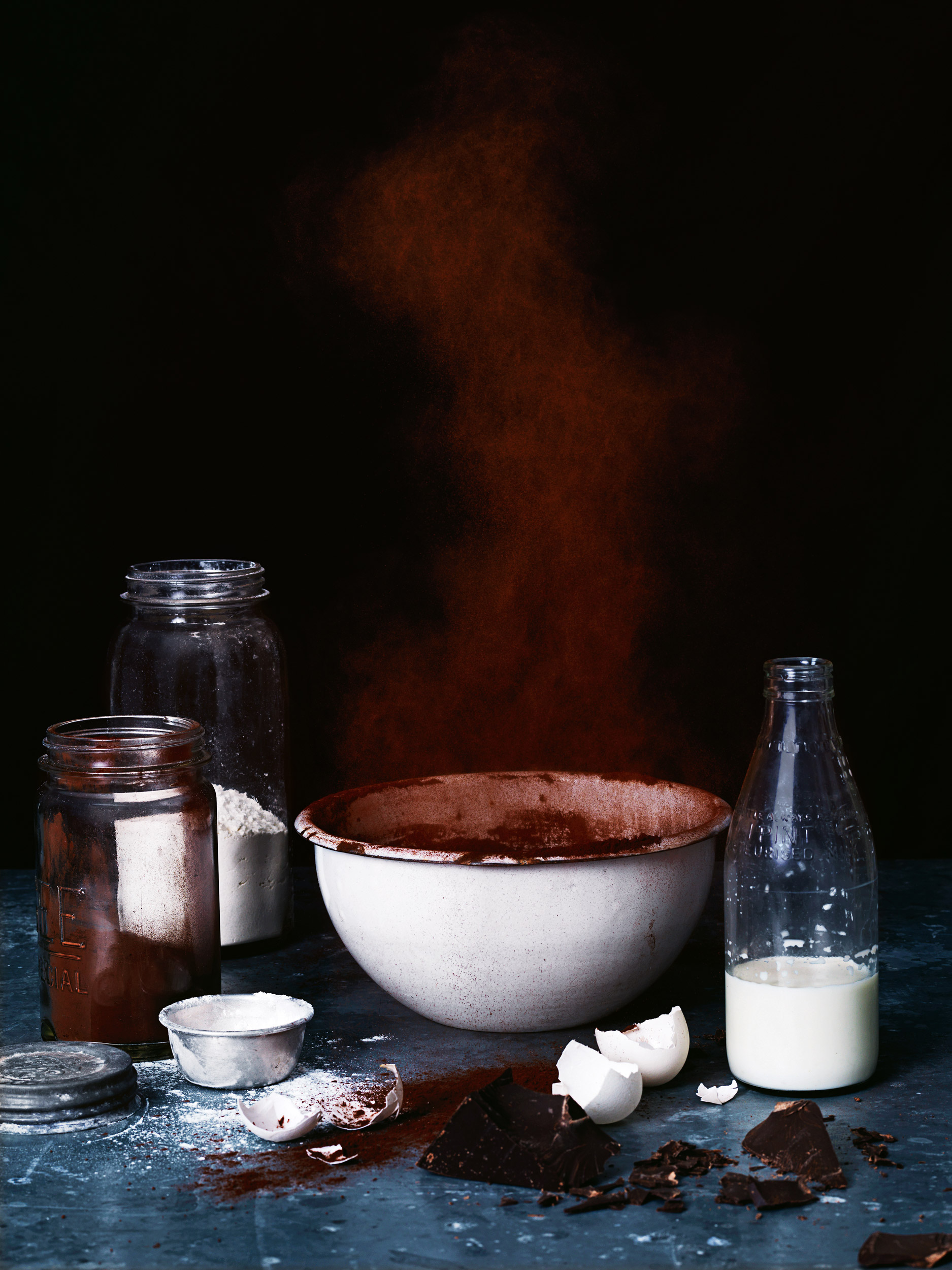 Still life shot of baking ingredients including a bowl, milk, flour, chocolate, and eggshells on a textured surface with dark backdrop.