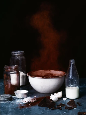 Still life shot of baking ingredients including a bowl, milk, flour, chocolate, and eggshells on a textured surface with dark backdrop.