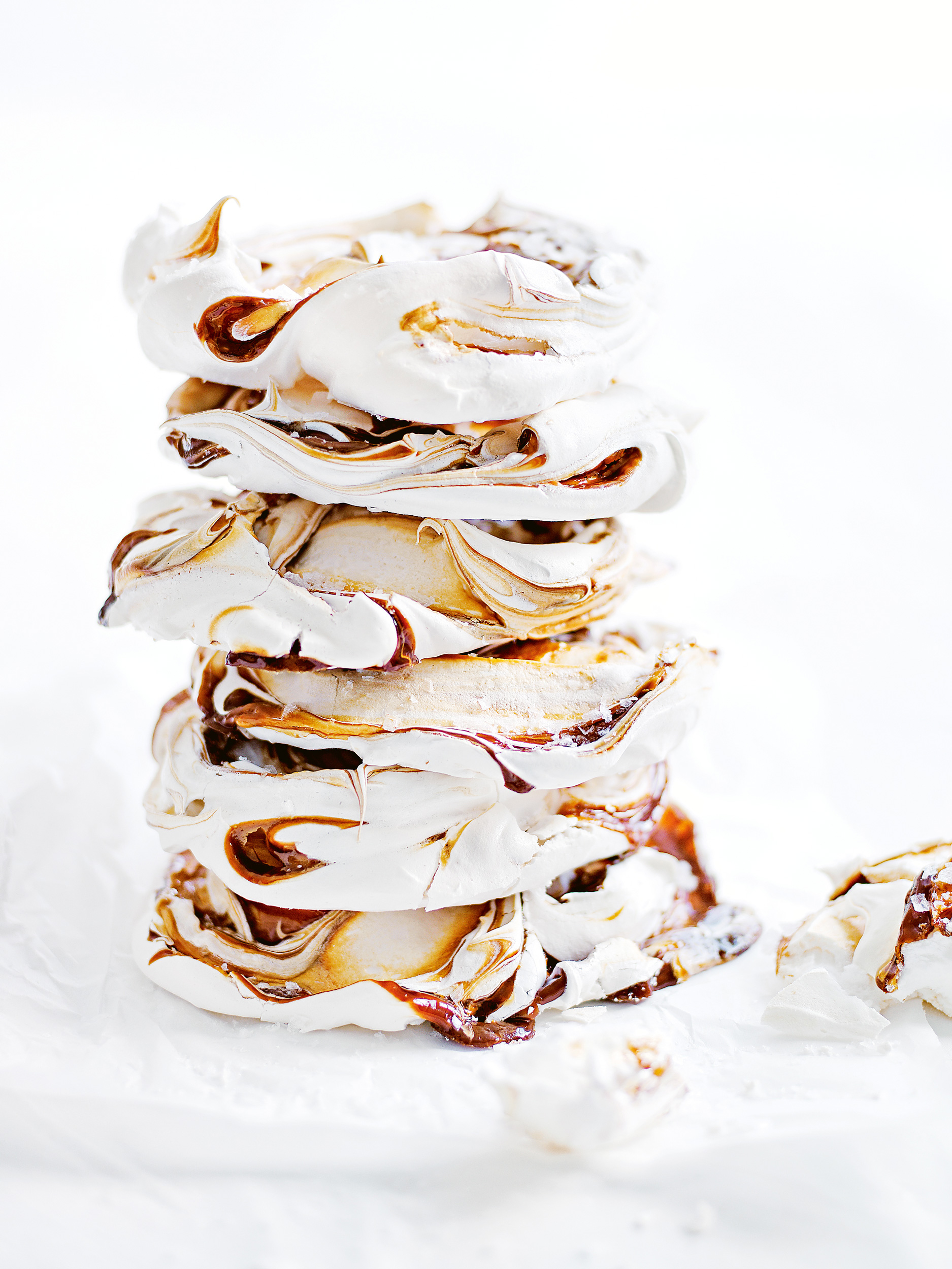 Stack of meringue cookies drizzled with chocolate on a white surface, showcasing the dessert's delicate texture and swirl design.