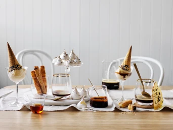 A styled arrangement of desserts, ice cream cones in glasses, pastries, and coffee on a wooden table with a white paneled wall background.