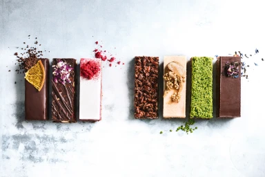 Seven gourmet chocolate and raw dessert bars arranged in a row on a weathered grey concrete surface, styled by Steve Pearce