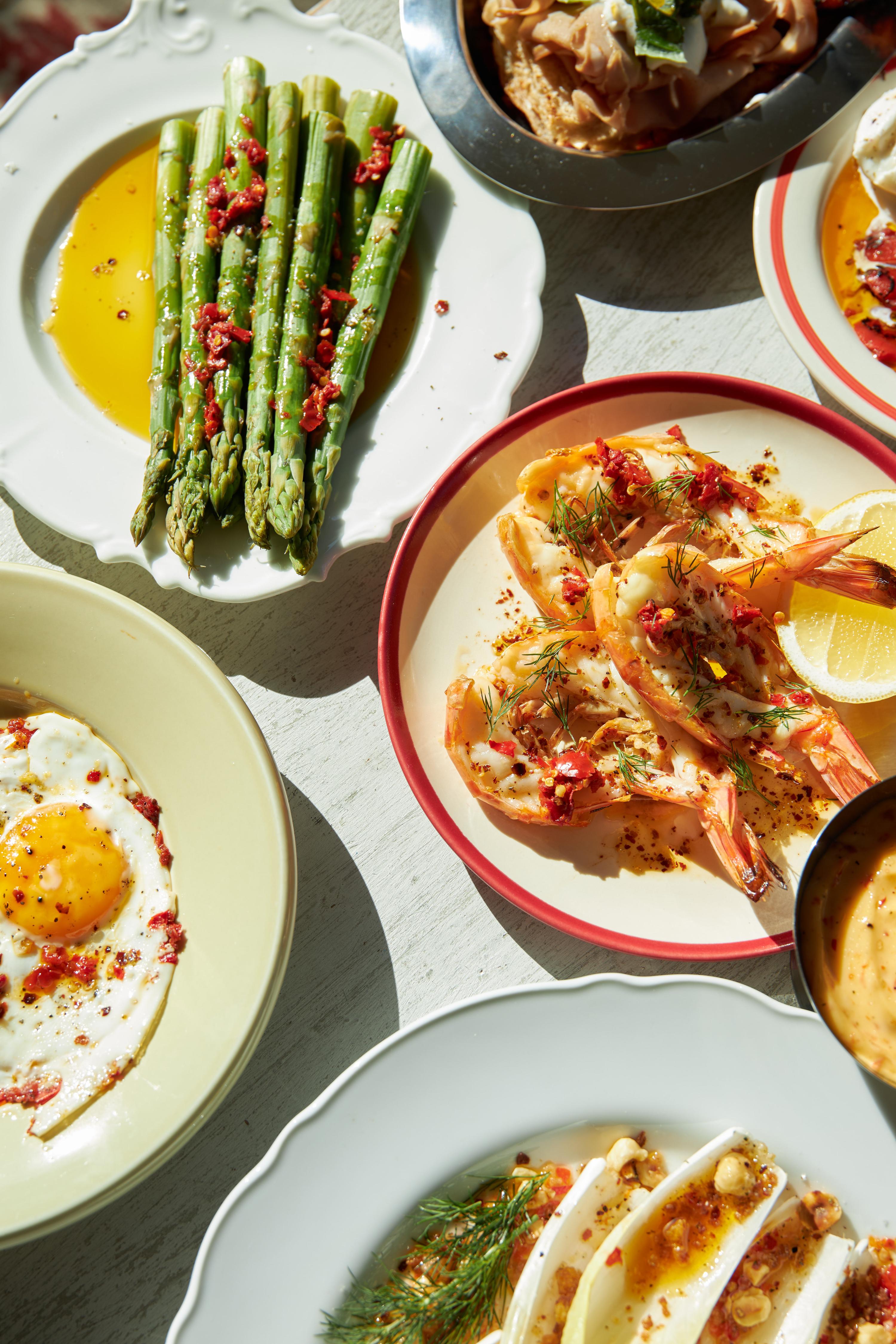 Overhead shot of a table with a variety of colourful dishes, including grilled shrimp, asparagus, fried eggs, and endive salad.