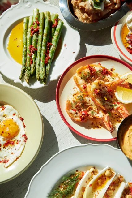 Overhead shot of a table with a variety of colourful dishes, including grilled shrimp, asparagus, fried eggs, and endive salad.