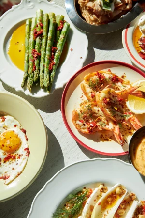 Overhead shot of a table with a variety of colourful dishes, including grilled shrimp, asparagus, fried eggs, and endive salad.