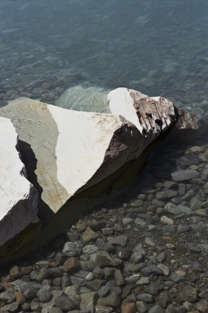 Large white rocks sit in clear water, smaller stones visible beneath the surface of the water in an outdoor setting.