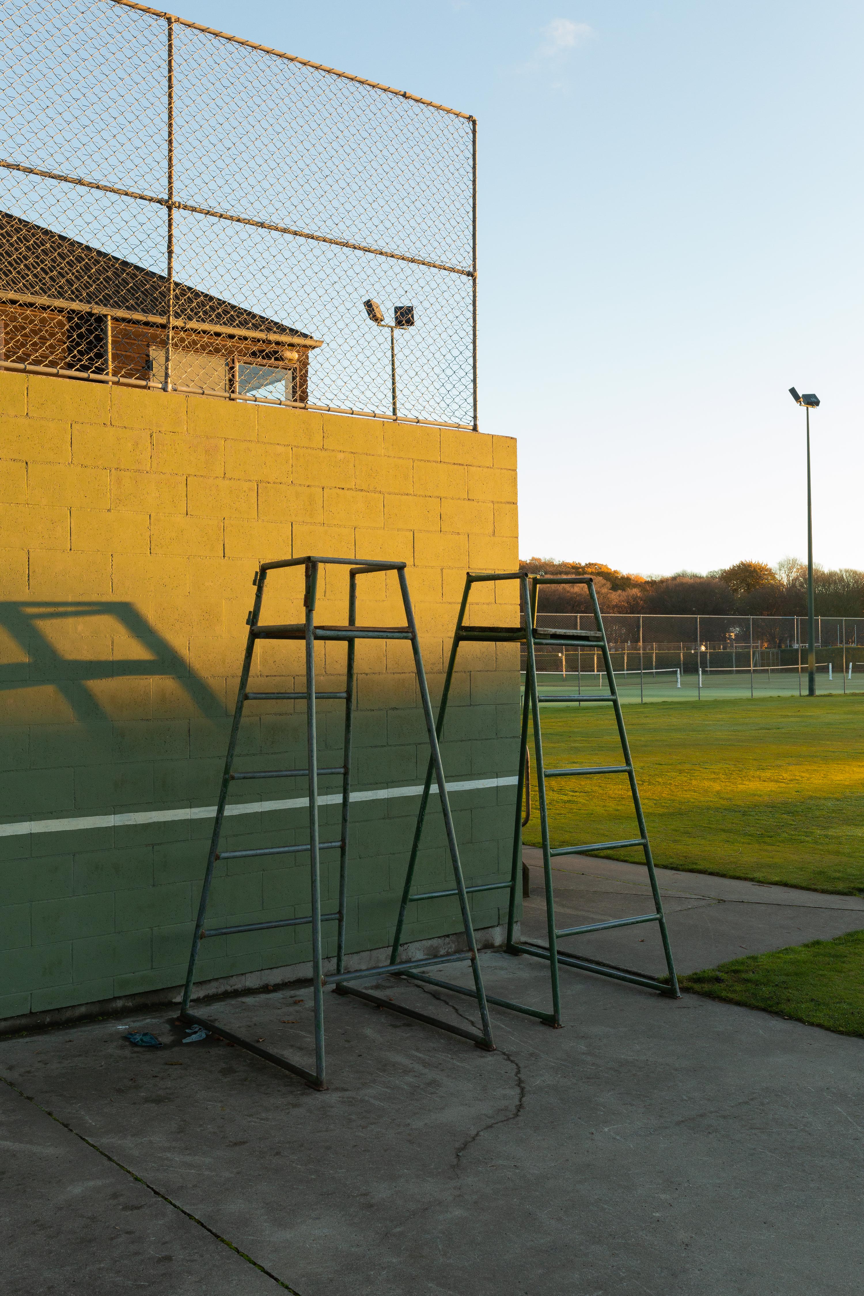 Two tennis umpire chairs stand on a green court near a yellow brick wall, bathed in the warm glow of golden hour light.