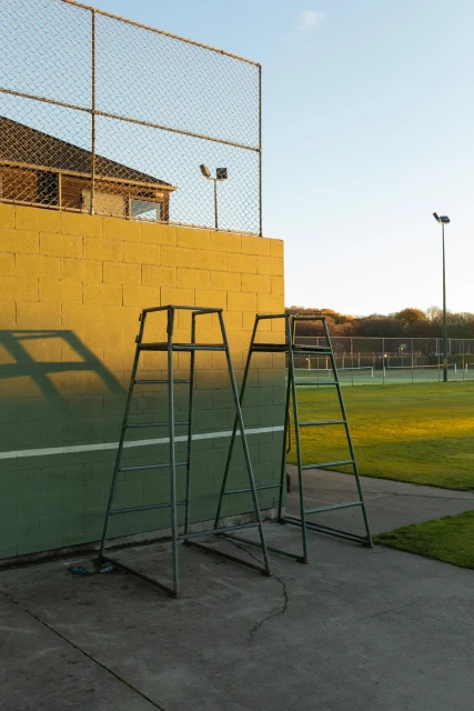 Two tennis umpire chairs stand on a green court near a yellow brick wall, bathed in the warm glow of golden hour light.