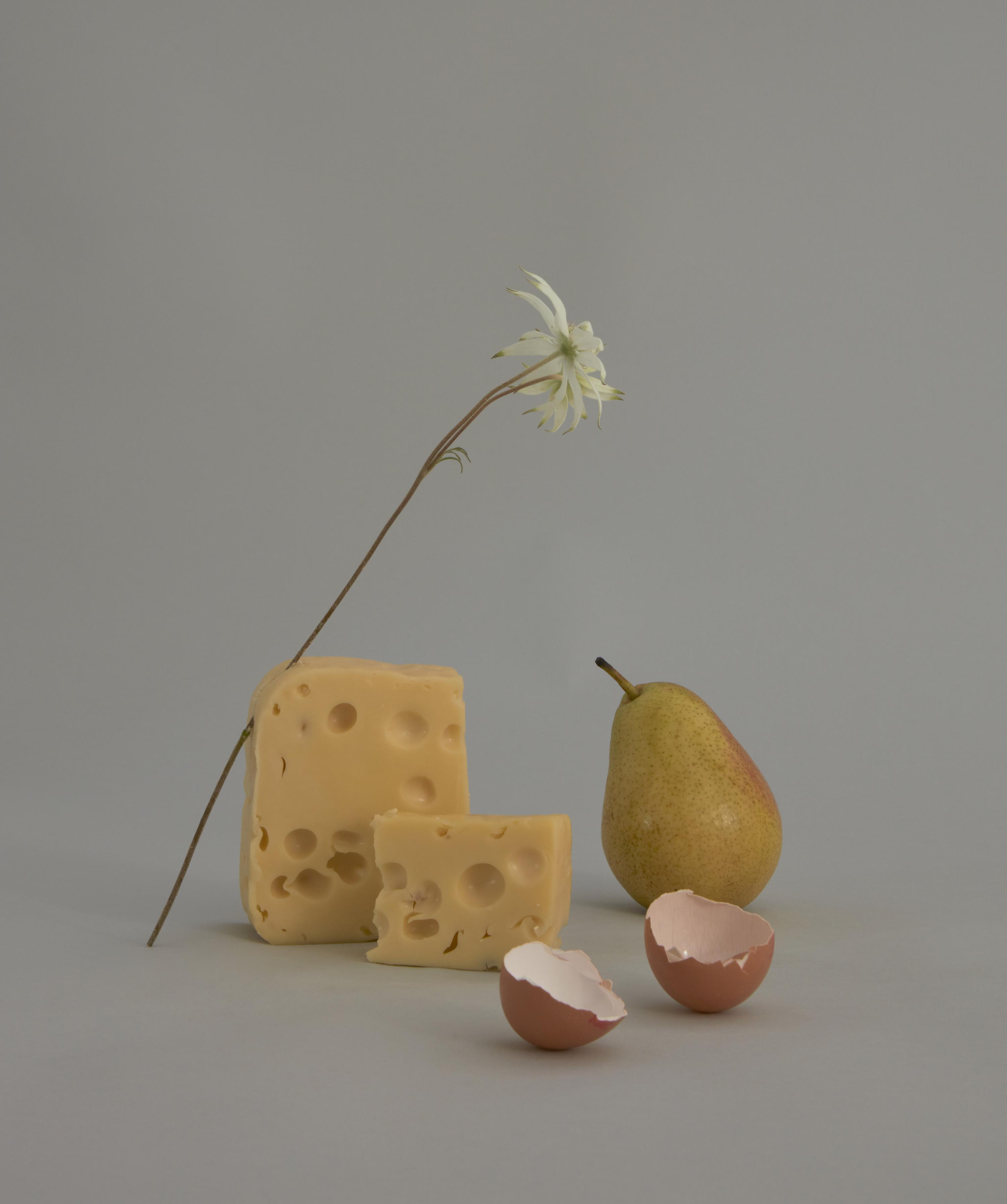 Still life shot featuring cheese, a pear, a flower, and eggshells against a neutral gray backdrop, capturing a simple and elegant composition.