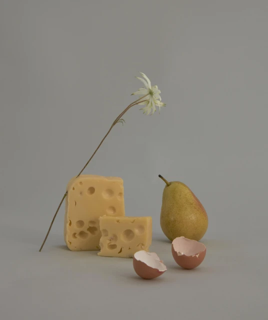 Still life shot featuring cheese, a pear, a flower, and eggshells against a neutral gray backdrop, capturing a simple and elegant composition.