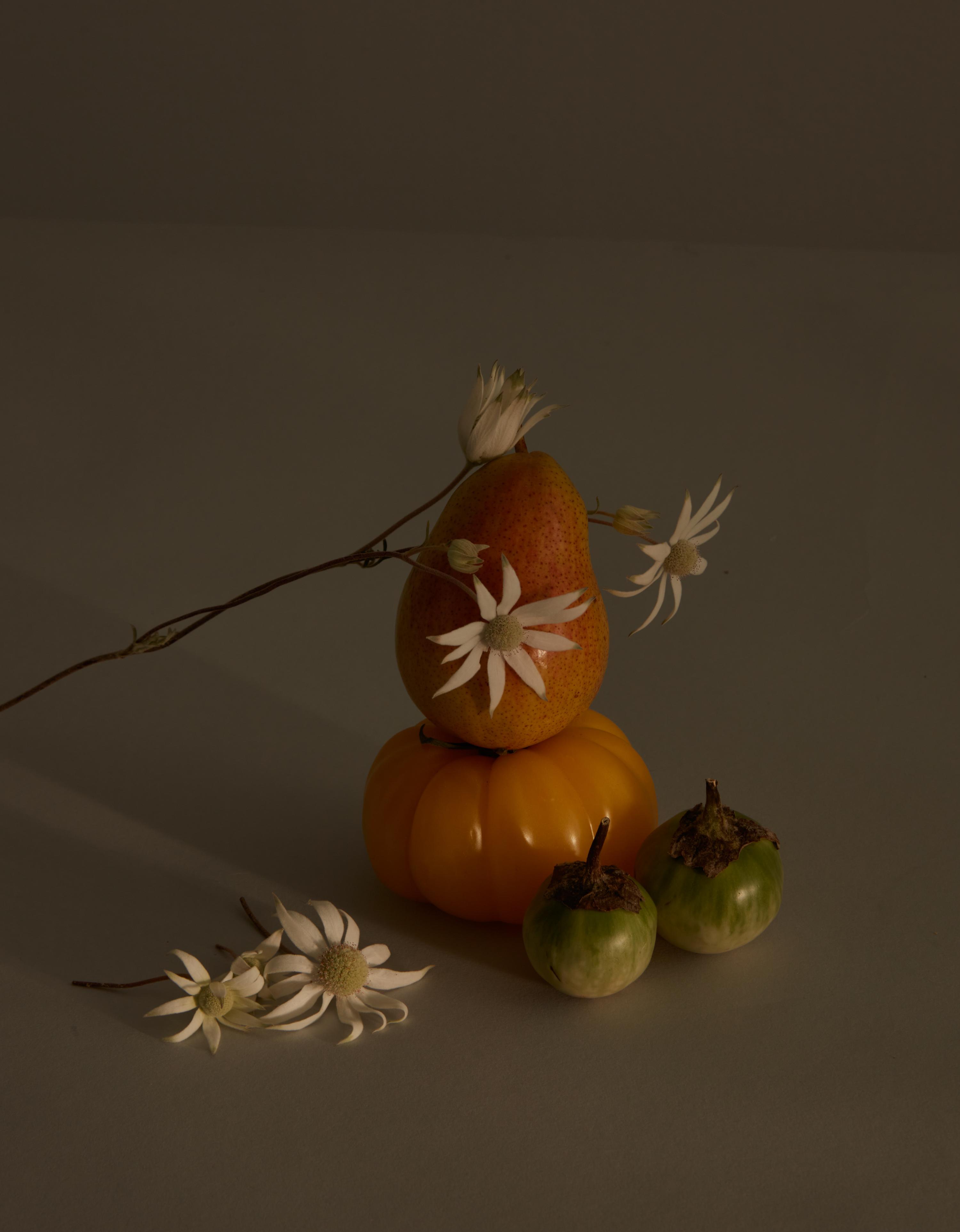 Still life photograph of a pear atop a pumpkin, adorned with white flowers, accompanied by two green eggplants on a neutral surface.